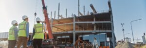Three construction workers in safety gear observe a building under construction with cranes and equipment on site.