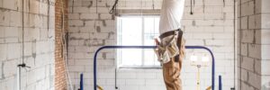 A construction worker in safety gear drills into the ceiling while standing on a scaffold in a partially finished room.