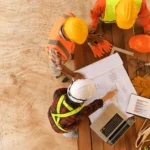 Three construction workers review blueprints and charts on a wooden table with safety gear and a laptop.