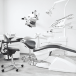 Modern dental office with a reclining dental chair, overhead light, equipment, and a stool on a clean, light-colored floor.