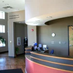 Reception area of a medical office with a curved desk, computer, wall clock, and a large vase of flowers on the counter.