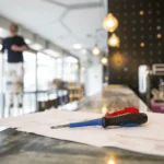 A screwdriver and building plans rest on a countertop with a worker on a ladder in the background.