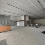 Large unfinished interior space with exposed ceiling, scattered furniture, ladders, and construction materials on the floor.