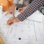 Two people review architectural blueprints on a desk with hard hats, a notebook, and measuring tools nearby.