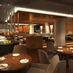 Modern restaurant interior with round wooden tables, set for dining, upholstered chairs, and warm ambient lighting.