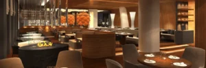 Modern restaurant interior with round wooden tables, set for dining, upholstered chairs, and warm ambient lighting.