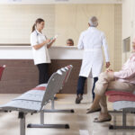 An older woman sits in a waiting area while two doctors talk at a reception desk in a medical office.
